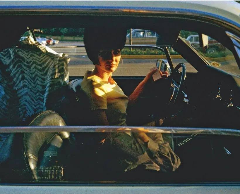 Colour photograph of a woman sat in the driver's seat of a car with clothes hung up behind her.