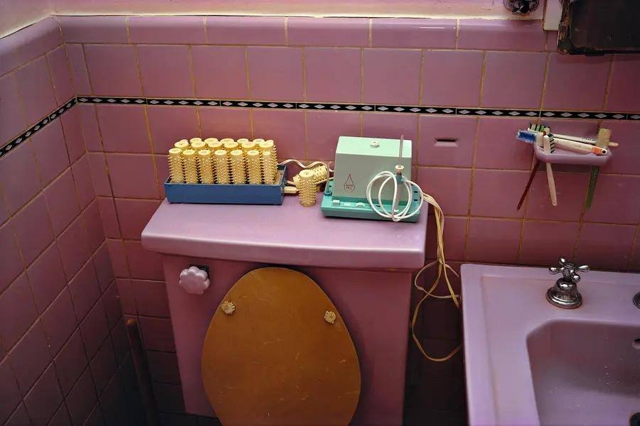 Colour photograph of a pink bathroom, including a pink bathtub, pink walls and a pink toilet, with hair rollers on the cistern.
