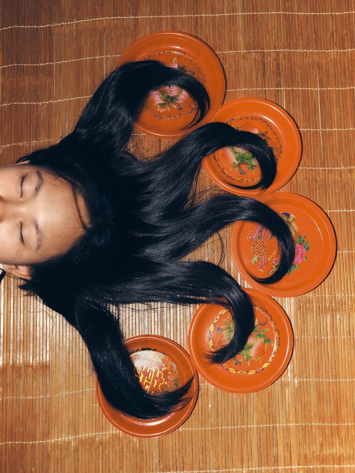 A woman with eyes closed lying on a woven mat, her long black hair combed in waves across five orange lacquered plates.
