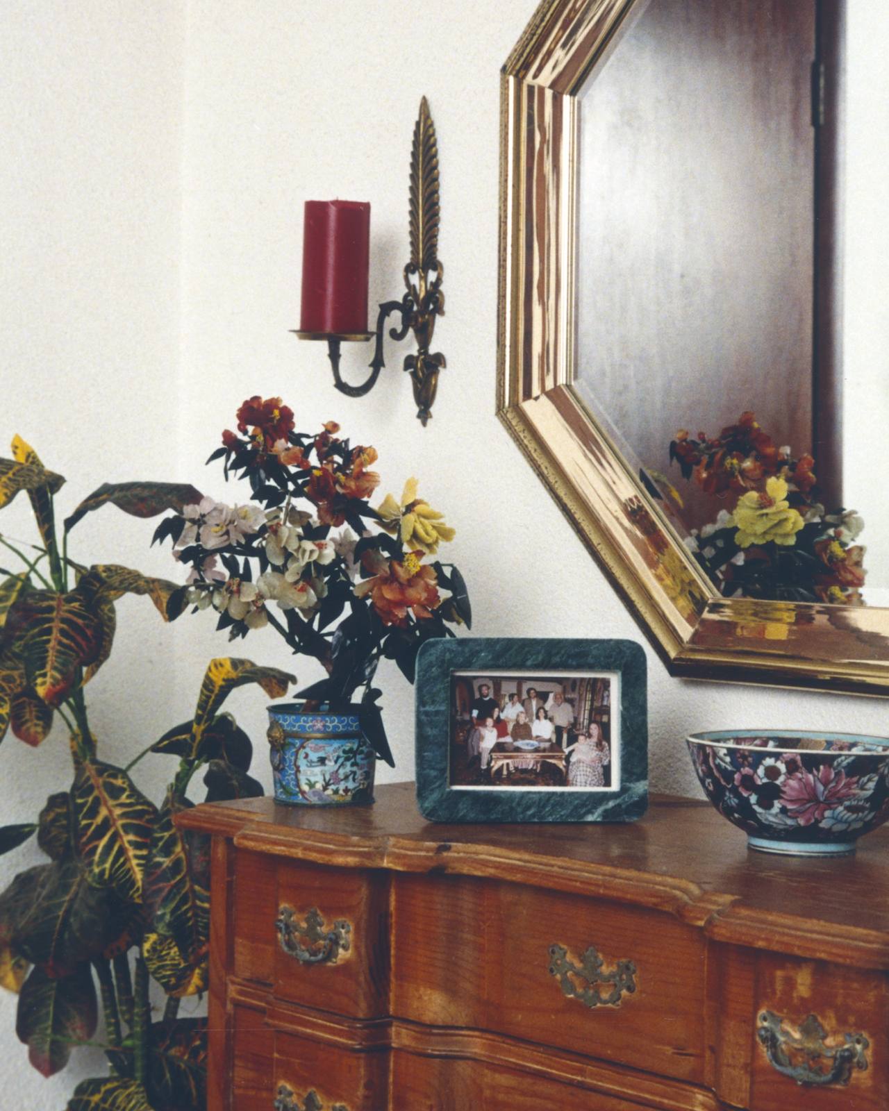 A domestic interior with a framed family photograph on a wooden dresser, beside a flowering plant and a painted bowl, below a mirror.