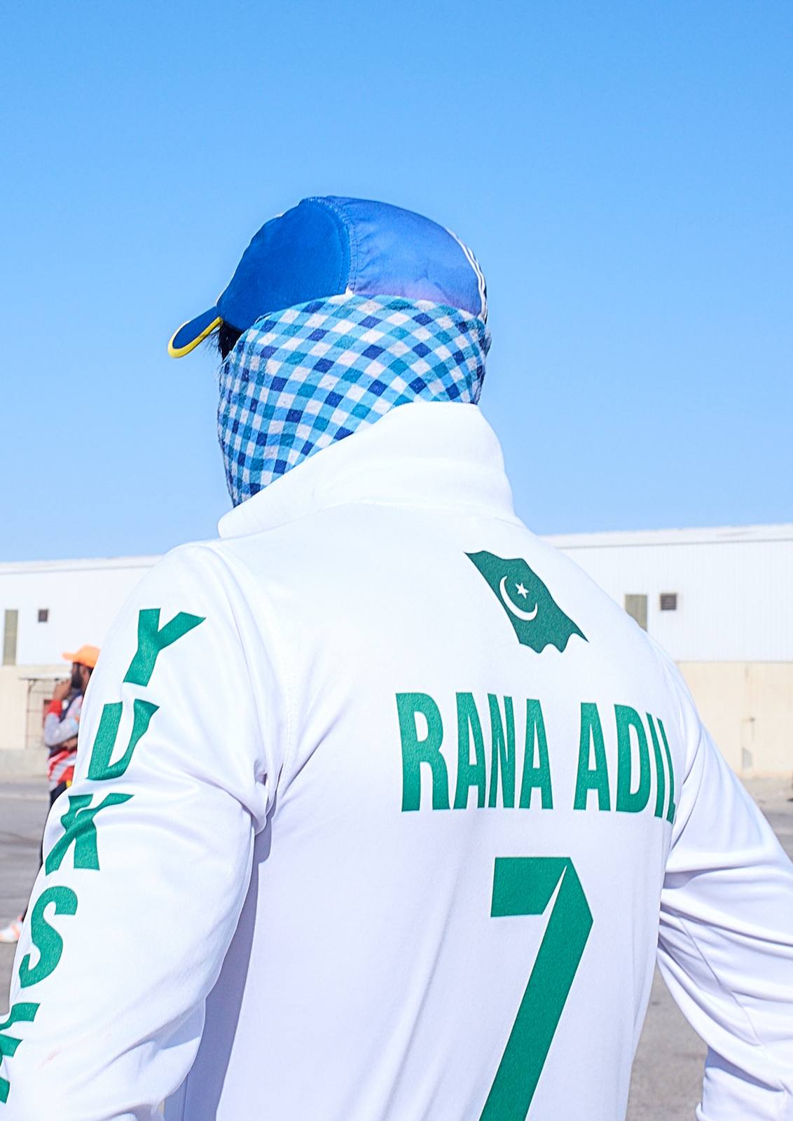 The back of a cricketer in a white shirt with a name and the number 7 in green, his head wrapped in checked cloth, against a blue sky.