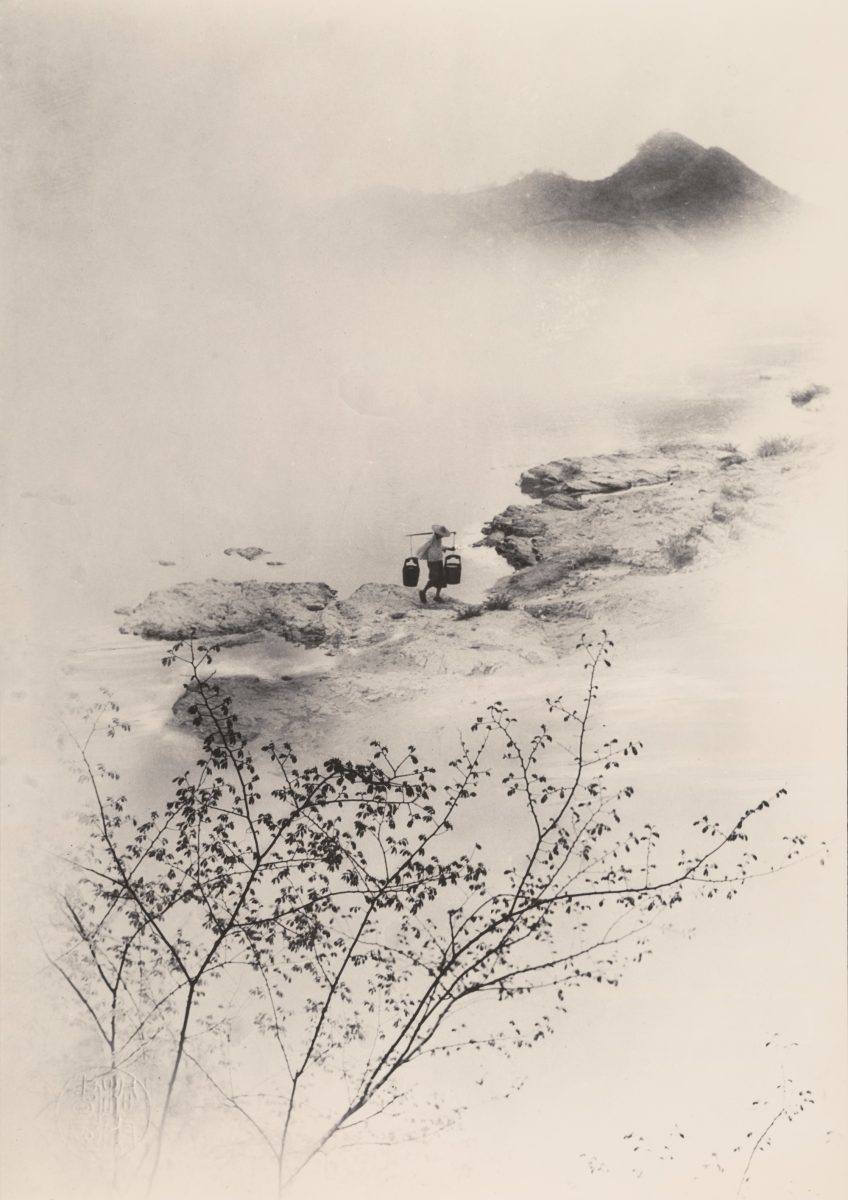 Composite photograph in the manner of Chinese ink painting: misty peaks, a figure carrying buckets on a rocky shore, blossoming branches, a red seal.