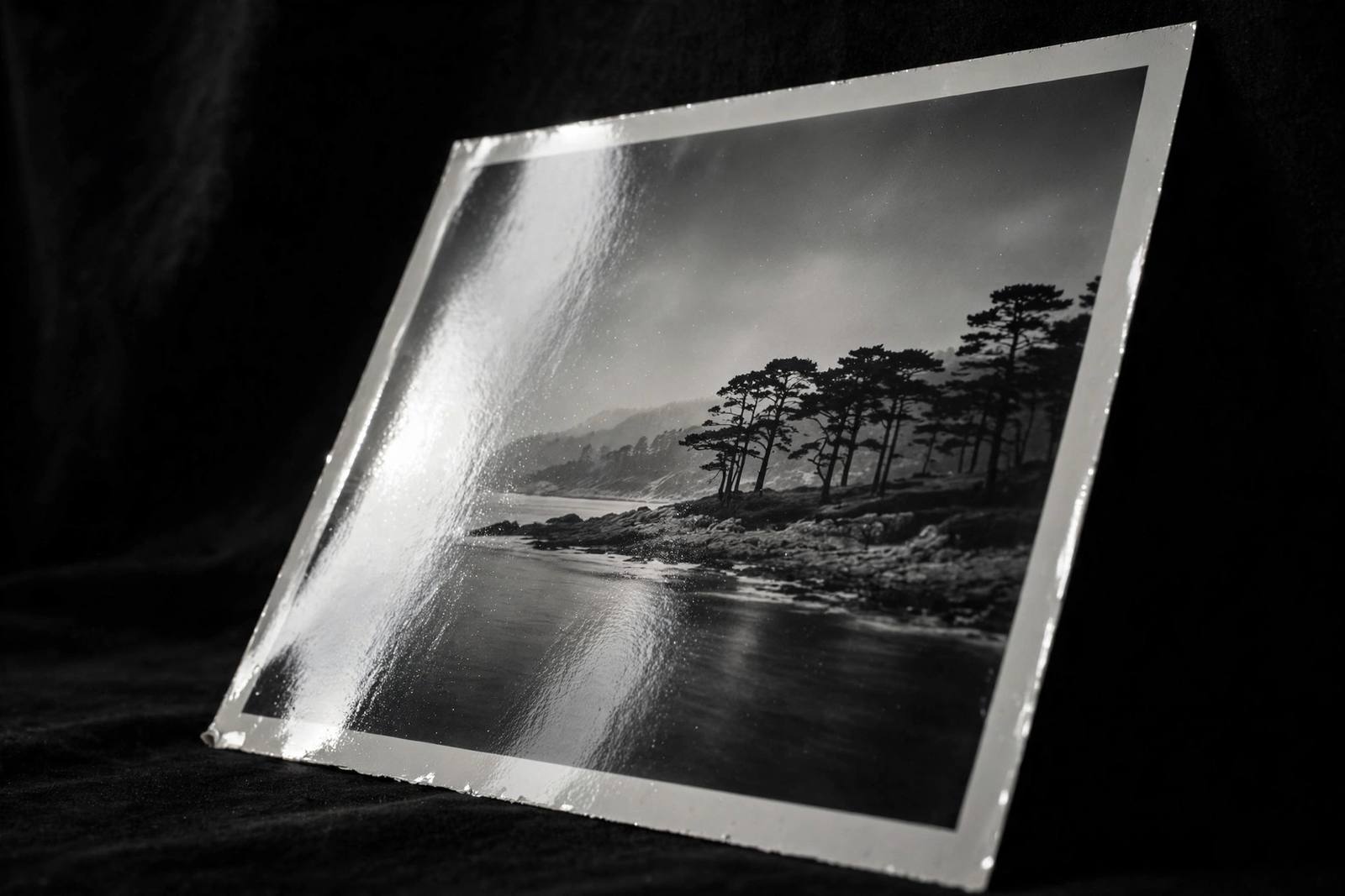 A black and white photograph held at a steep angle, its glossy emulsion surface catching the light in a visible sheen.