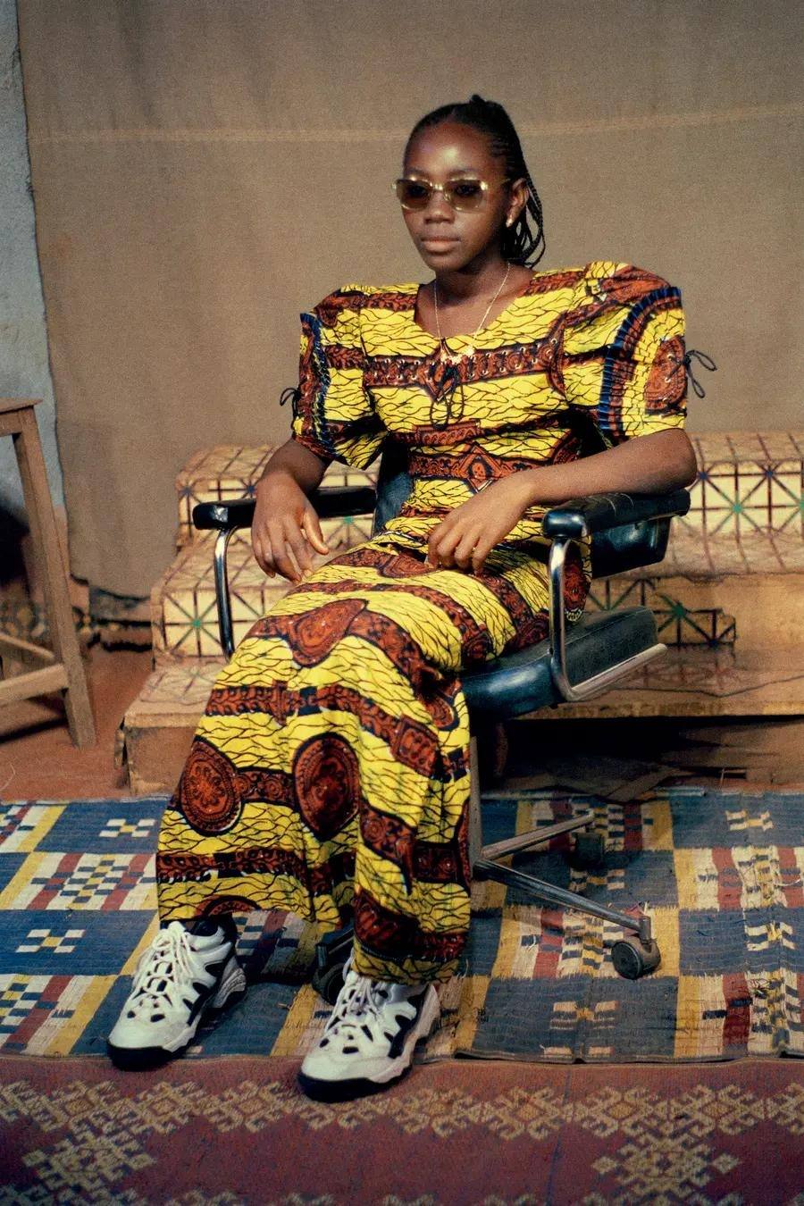 Colour photograph of a woman sat on a chair. She wears a traditional dress with sunglasses, and trainers.