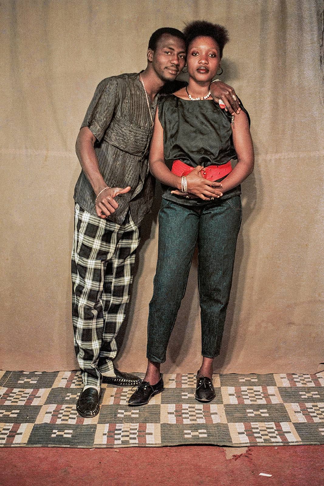 Colour studio portrait of a couple standing on a patterned mat, he in checked trousers, she in a black top, jeans and a wide red belt.