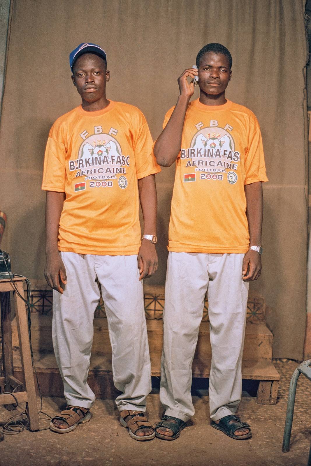 Colour studio portrait of two young men in matching orange Burkina Faso football shirts, one holding a mobile phone to his ear.