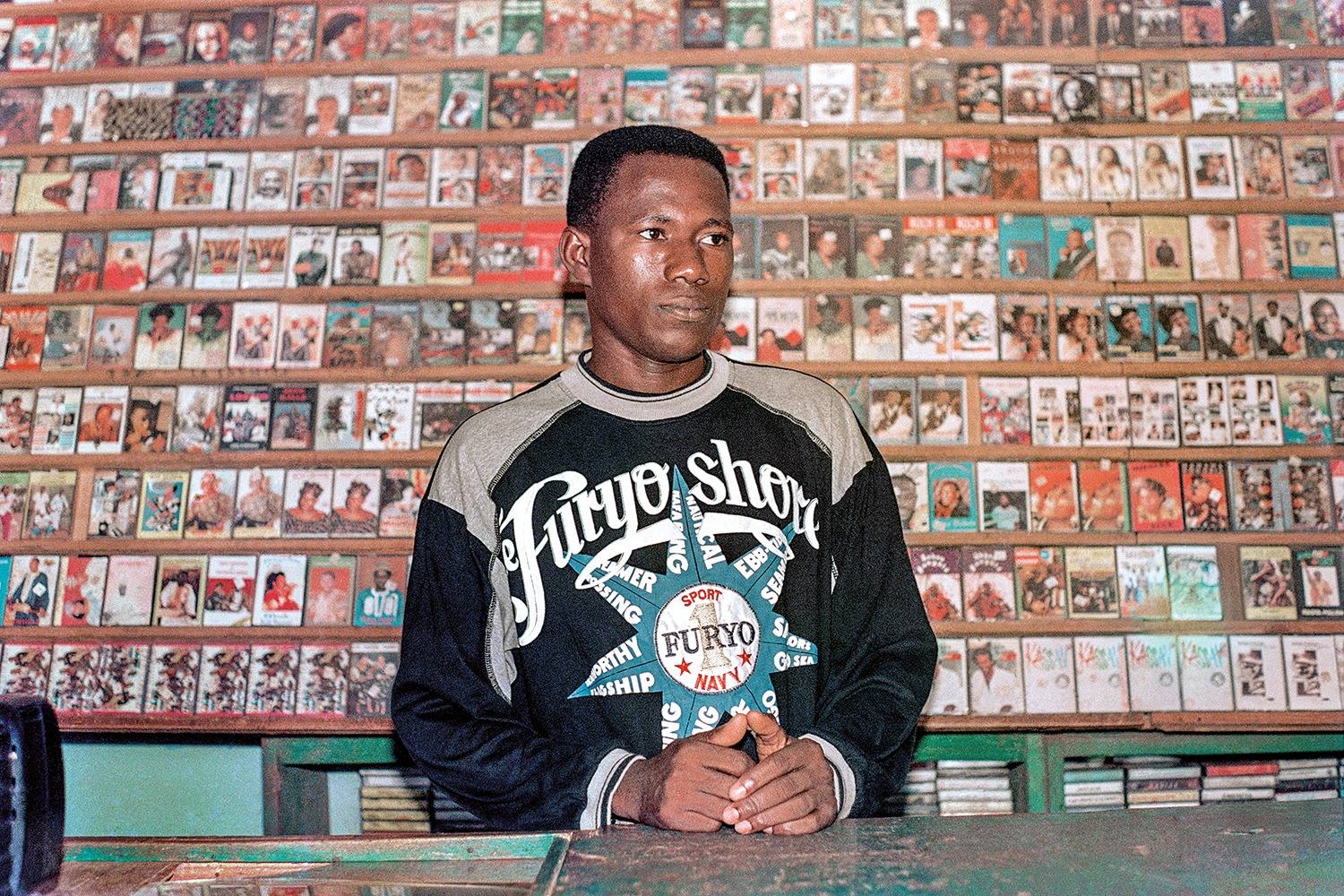 Colour photograph of a man standing behind a shop counter, hands folded, before shelves covered with cassette and video covers.