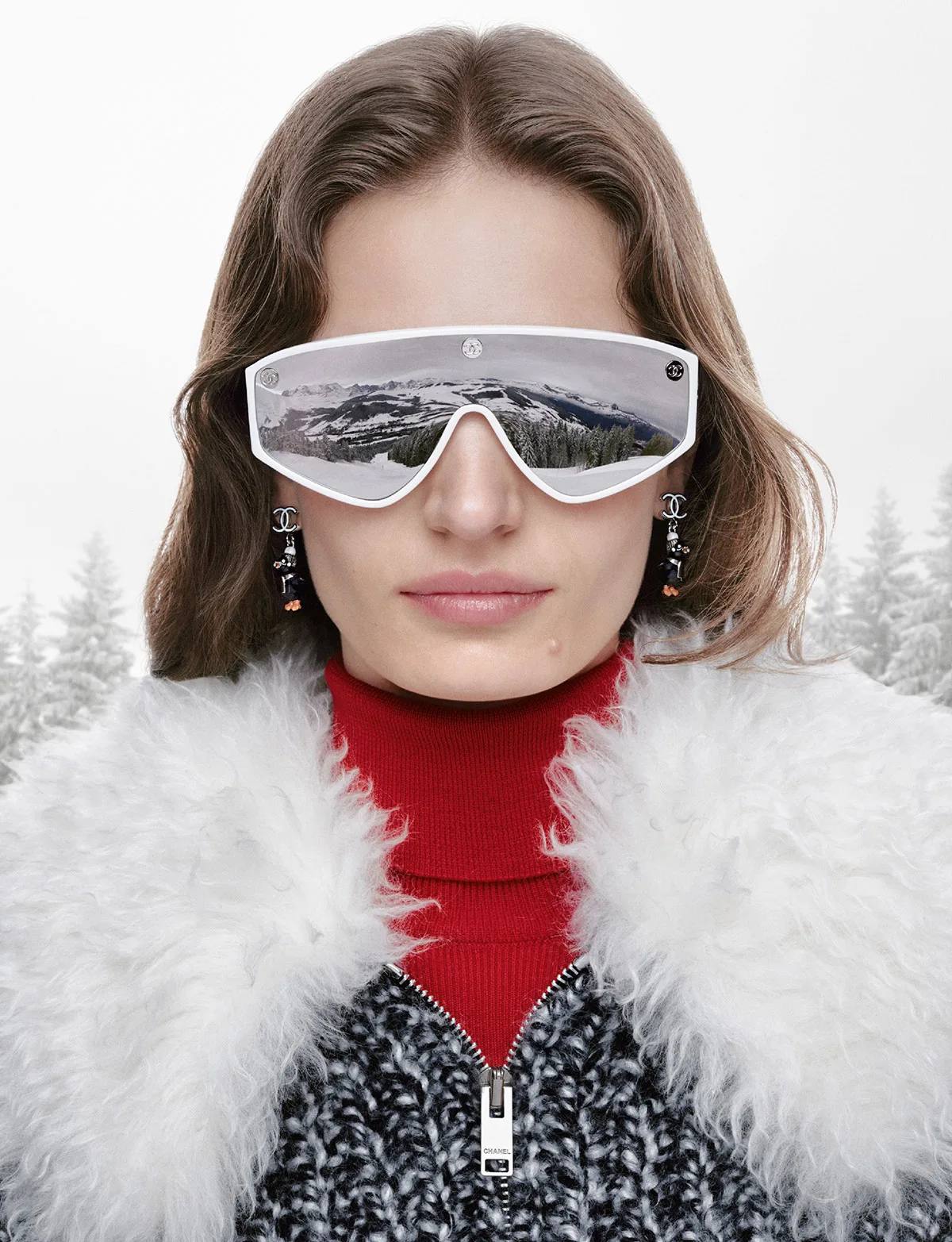 Close portrait from the Chanel Coco Neige 2026 campaign, a model in mirrored shield sunglasses reflecting a snowy mountain, red turtleneck and white shaggy collar, penguin earrings visible